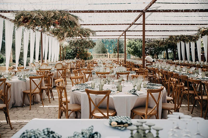Banquete de boda montado al aire libre bajo pérgola, mesas vestidas con mantelería y sillas de madera