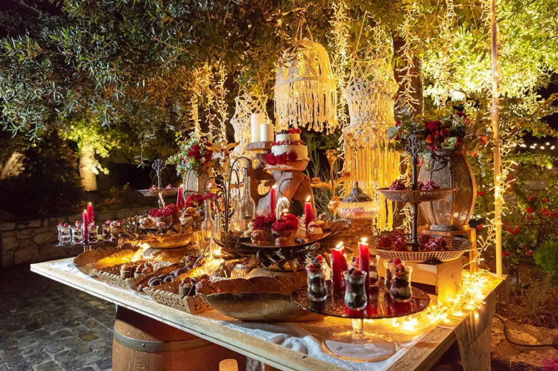 Mesa de postres y dulces iluminada al aire libre, velas, flores y luces de guirnalda entre olivos